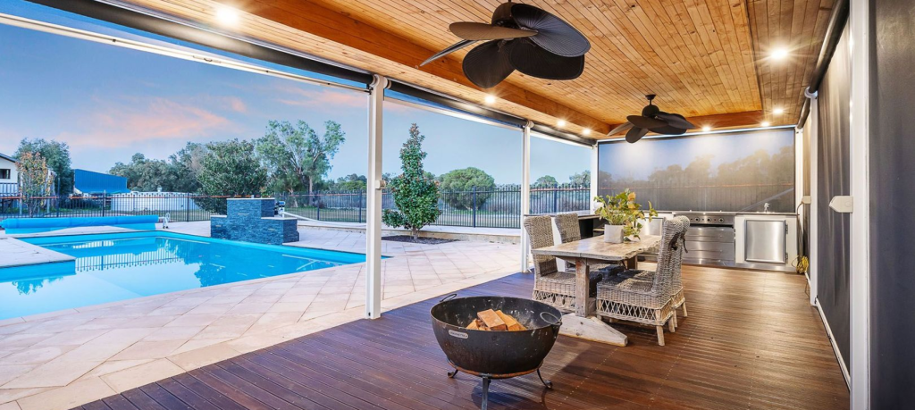 Outdoor living area that has a feature ceiling recess lined with timber panelling. A swimming pool, outdoor kitchen, and trees can be seen in the background.